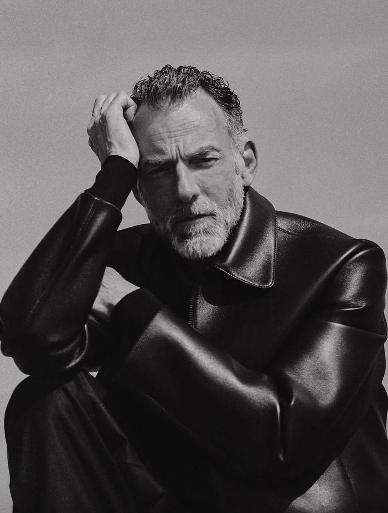 Monochrome portrait of a mature adult male seated indoors wearing a black leather jacket and trousers against a neutral background.
