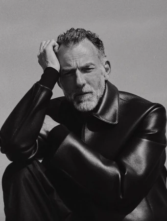 Monochrome portrait of a mature adult male seated indoors wearing a black leather jacket and trousers against a neutral background.