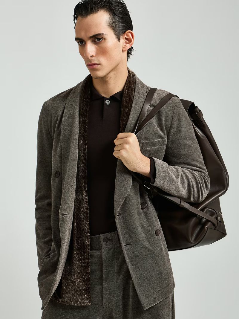 A stylish man in a brown suit and polo shirt with a brown shoulder bag poses in a photo studio with neutral lighting.
