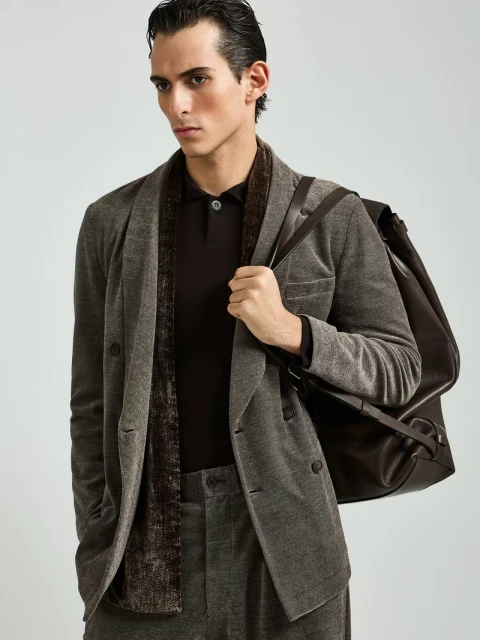A stylish man in a brown suit and polo shirt with a brown shoulder bag poses in a photo studio with neutral lighting.