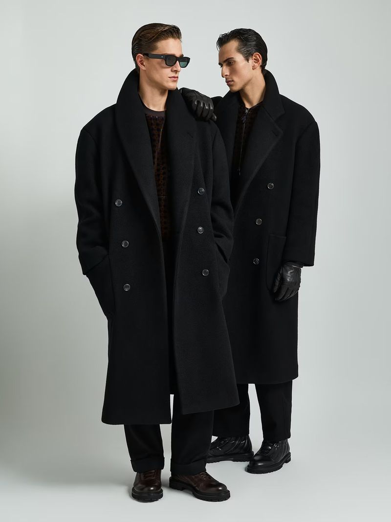 Two adult men in oversized black overcoats pose in a studio. One wears sunglasses and has a hand on the other's shoulder in an indoor setting.