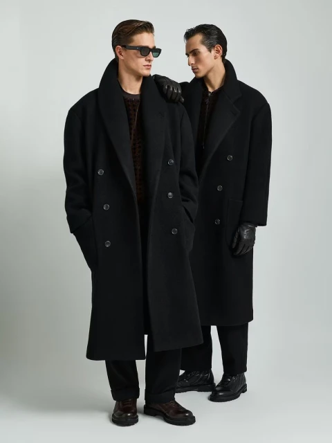 Two adult men in oversized black overcoats pose in a studio. One wears sunglasses and has a hand on the other's shoulder in an indoor setting.