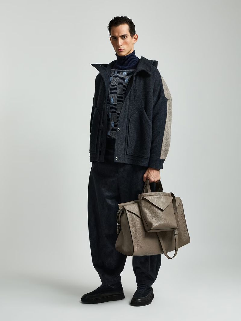 Full-length shot of a male model wearing a navy turtleneck and jacket while holding a leather bag on a neutral background.
