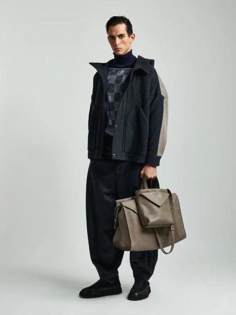 Full-length shot of a male model wearing a navy turtleneck and jacket while holding a leather bag on a neutral background.