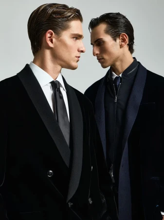 Two adult men in formal wear stand close together in a studio setting, one in a velvet tuxedo and the other in a dark suit and coat.