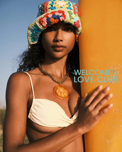 A woman in a crocheted hat and bikini top leans against a surfboard with ocean and sky background. Fashion, summer, beach, style.
