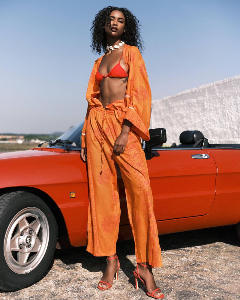 Elegant dark-skinned woman in orange outfit poses next to a red convertible car under a clear blue sky, exuding confidence and style.