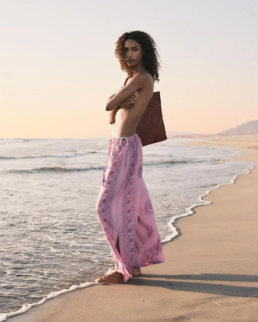 Woman with curly hair on a sandy beach at sunset wearing pink pants and carrying a dark-colored woven bag. Beachside fashion.