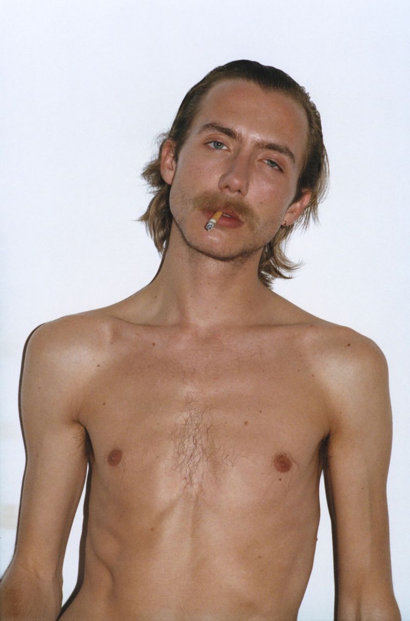 A shirtless adult male with blond hair and a mustache poses while smoking a cigarette in a brightly lit indoor environment against white backdrop.