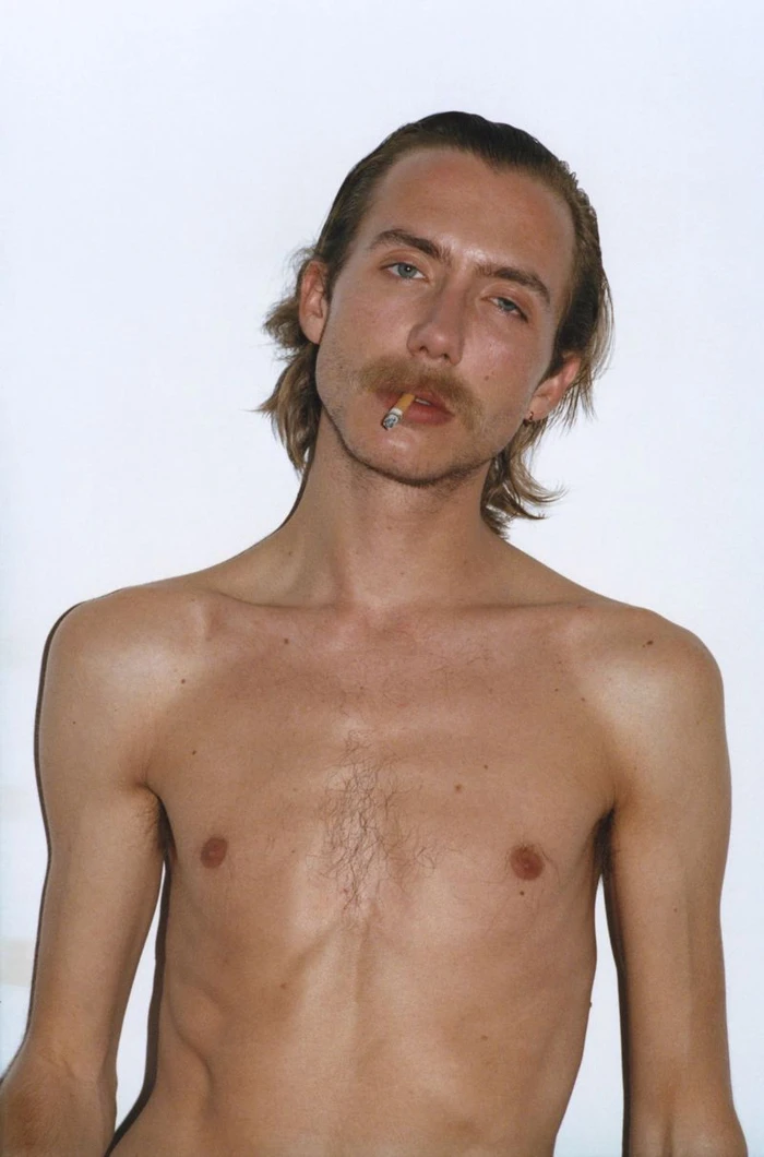 Portrait of a man with a mustache holding a cigarette in his mouth against a white seamless background.