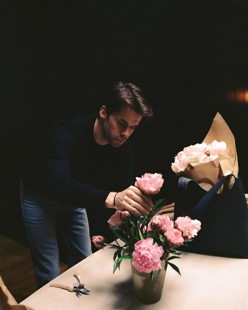 Adult male florist arranging pink peony flowers in a vase on a table with crafting tools indoors against a dark background.