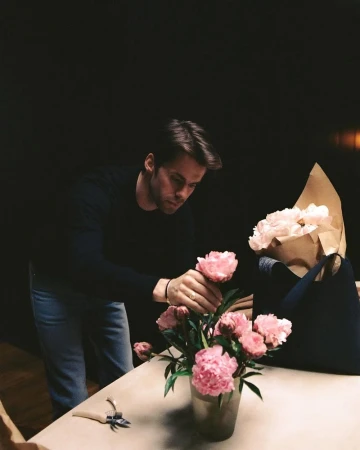 Adult male florist arranging pink peony flowers in a vase on a table with crafting tools indoors against a dark background.