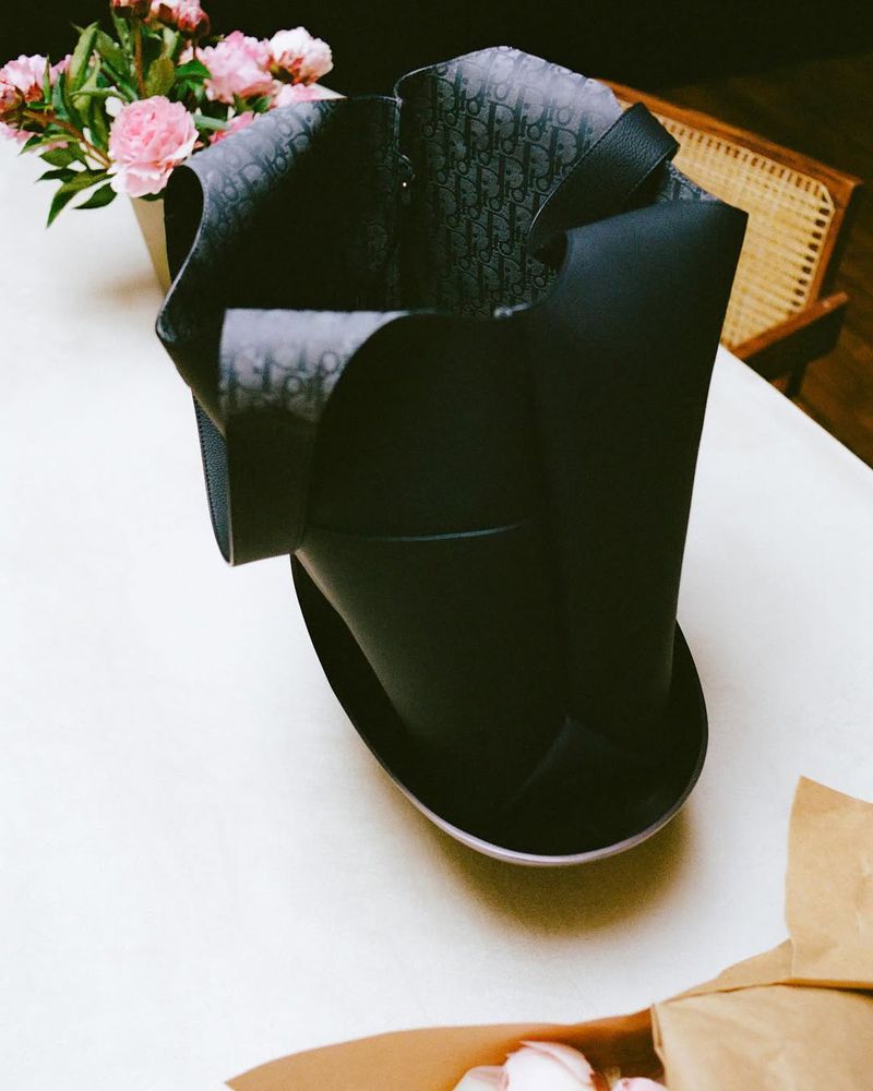 Elegant black tote bag with Dior monogram pattern on a white table next to pink flowers in a vase and a light wood chair.