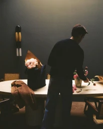 Man standing at a white oval table arranging flowers inside a moody dark-walled room for a Dior Men editorial project.