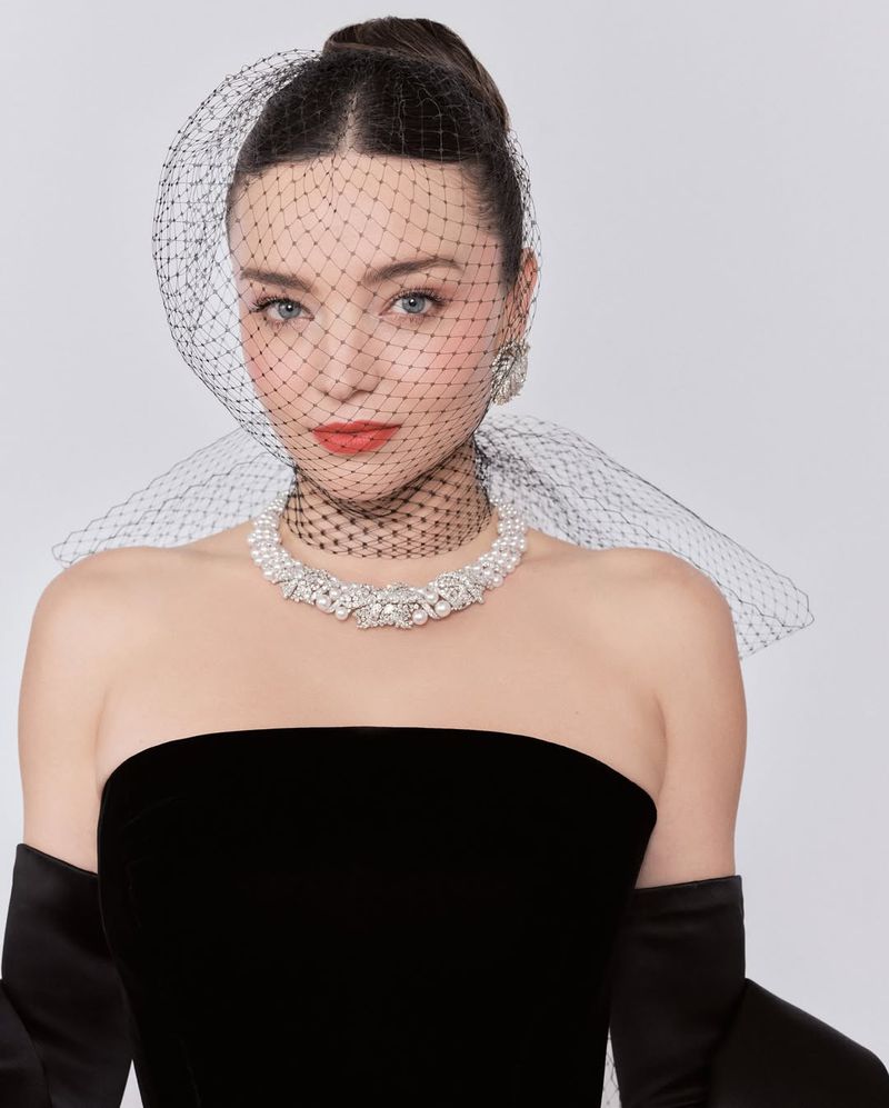 Elegant woman in black strapless dress, pearl necklace, and net veil against a white background in a portrait shot with jewelry.