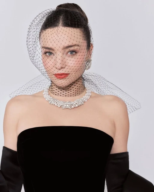 Elegant woman in black strapless dress, pearl necklace, and net veil against a white background in a portrait shot with jewelry.