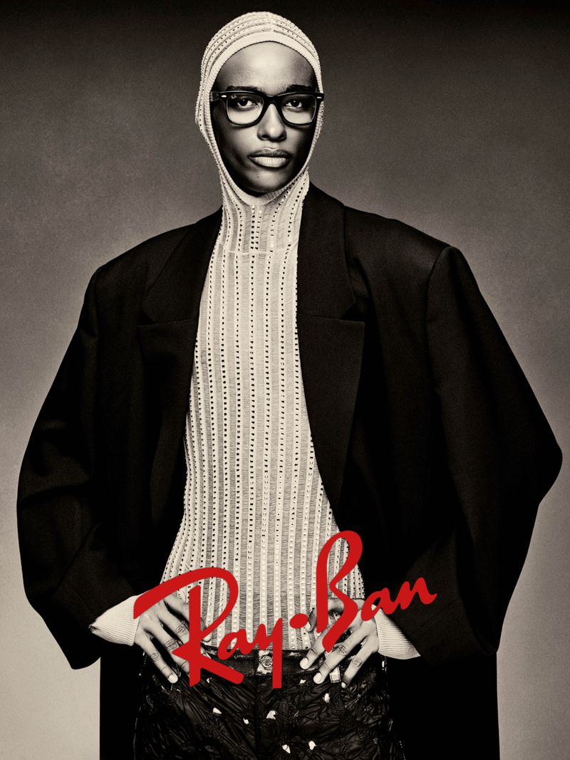 Fashionable Black model poses in a black blazer, glasses, and a textured top against a dark background in a studio portrait.
