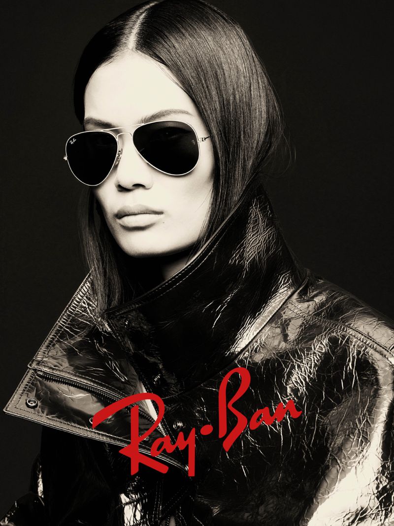 Woman with straight dark hair wearing aviator sunglasses and a shiny jacket. Ray-Ban logo displayed on lower part of the image.