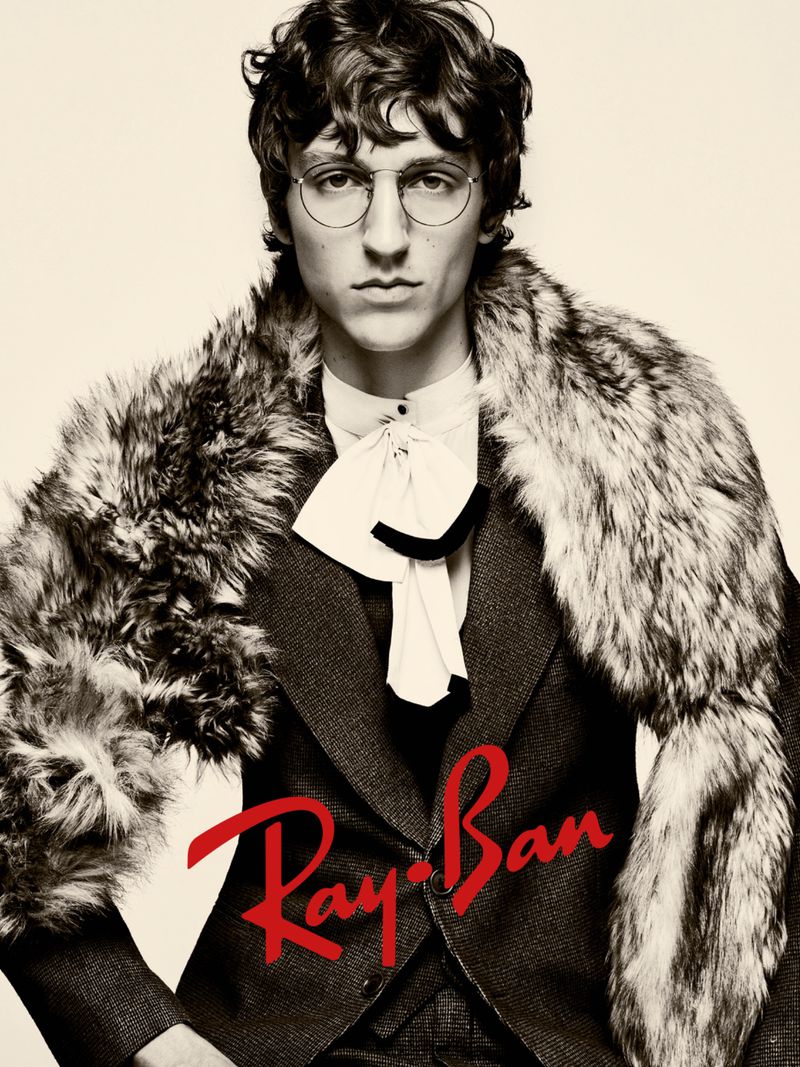 Portrait of a man wearing a tweed suit, a white ruffled shirt, round glasses, and a fur stole, with the Ray-Ban logo.