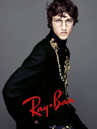 Fashion portrait of a young adult man in round eyeglasses, black turtleneck, and embellished blazer with Ray-Ban logo overlay.