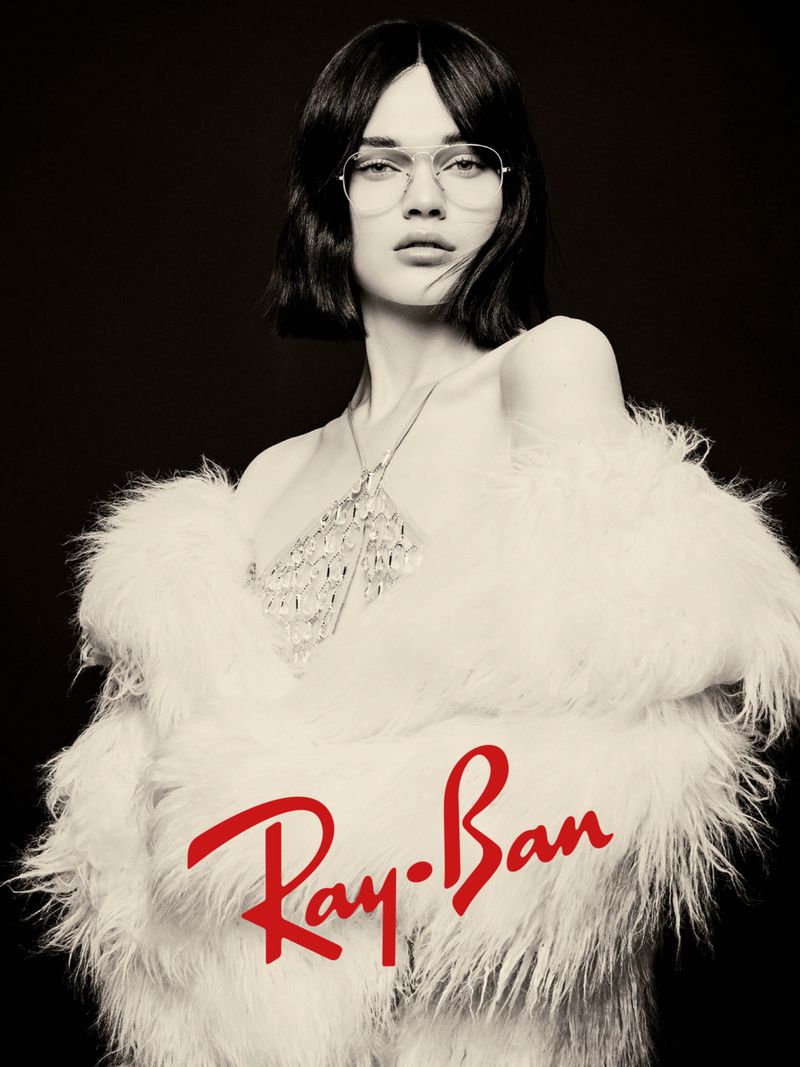 A woman wearing glasses and a white faux fur coat poses against a black background in a studio, showcasing a Ray-Ban logo.