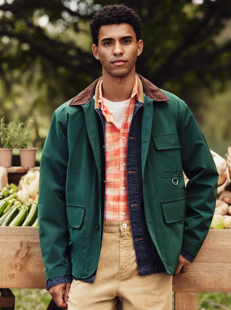A young adult man with an olive complexion stands outdoors wearing a green jacket and khaki pants against a backdrop of greenery.