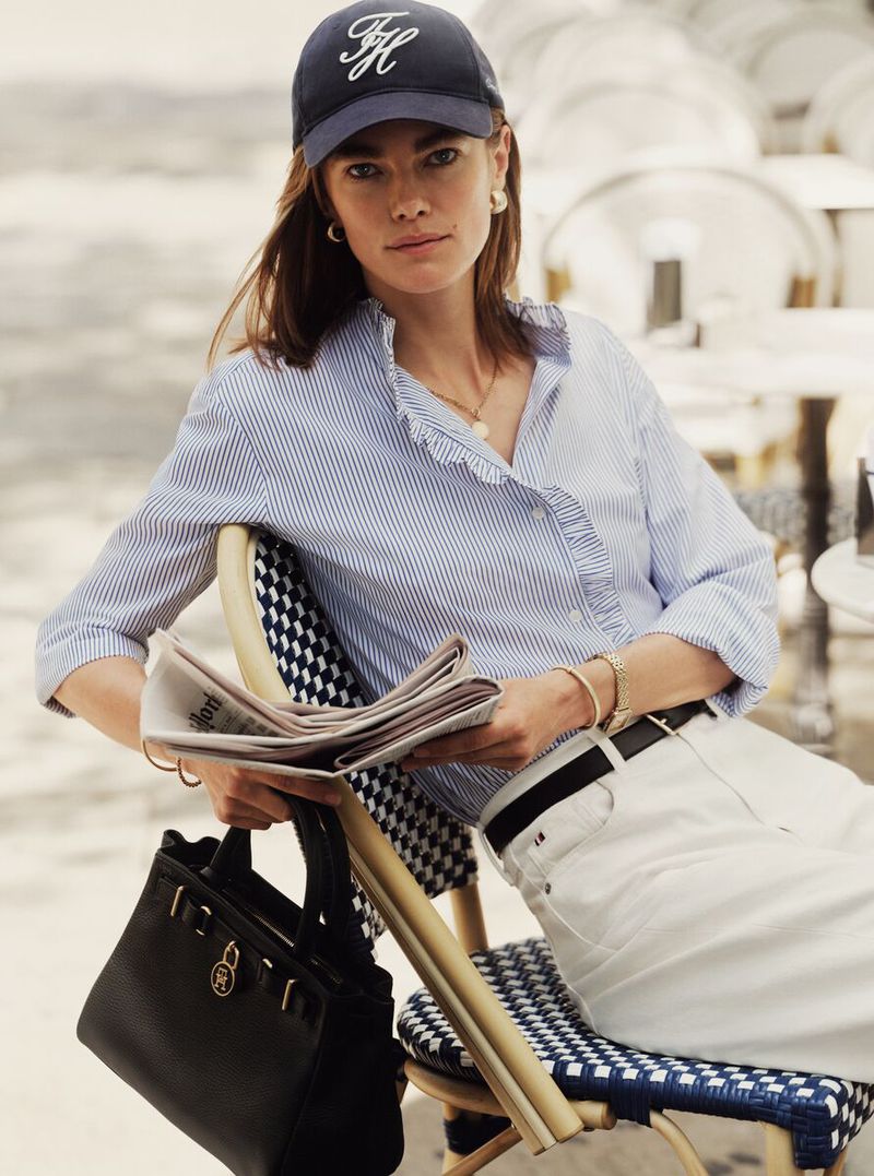 A woman in a blue and white striped shirt and baseball cap sitting on a chair with a newspaper and black purse in an outdoor setting.