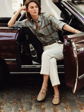 Adult woman sitting in a vintage convertible car wearing a plaid shirt, cream-colored slacks, and T-strap shoes outdoors on a cobblestone surface.