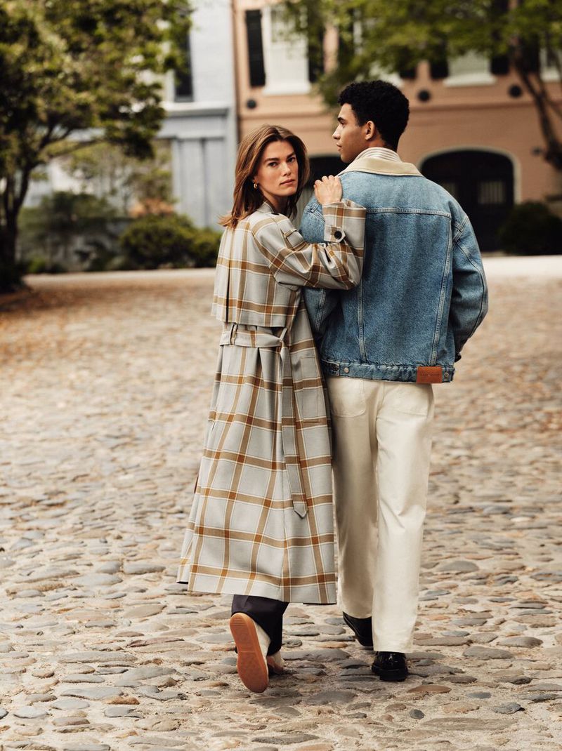 Couple walking on cobblestone street. Woman in plaid trench coat looks back at camera, man in denim jacket walks beside her.