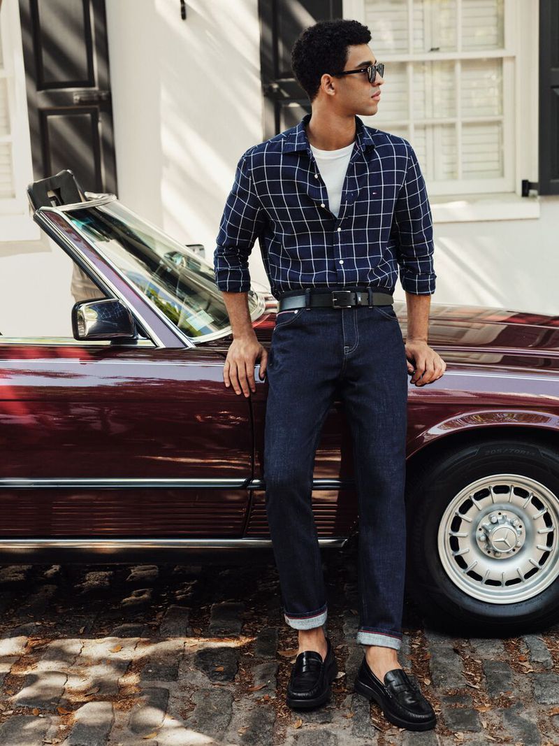 Stylish adult male poses next to a red convertible in casual-chic attire, featuring a plaid shirt, jeans, and loafers for a refined, relaxed look.