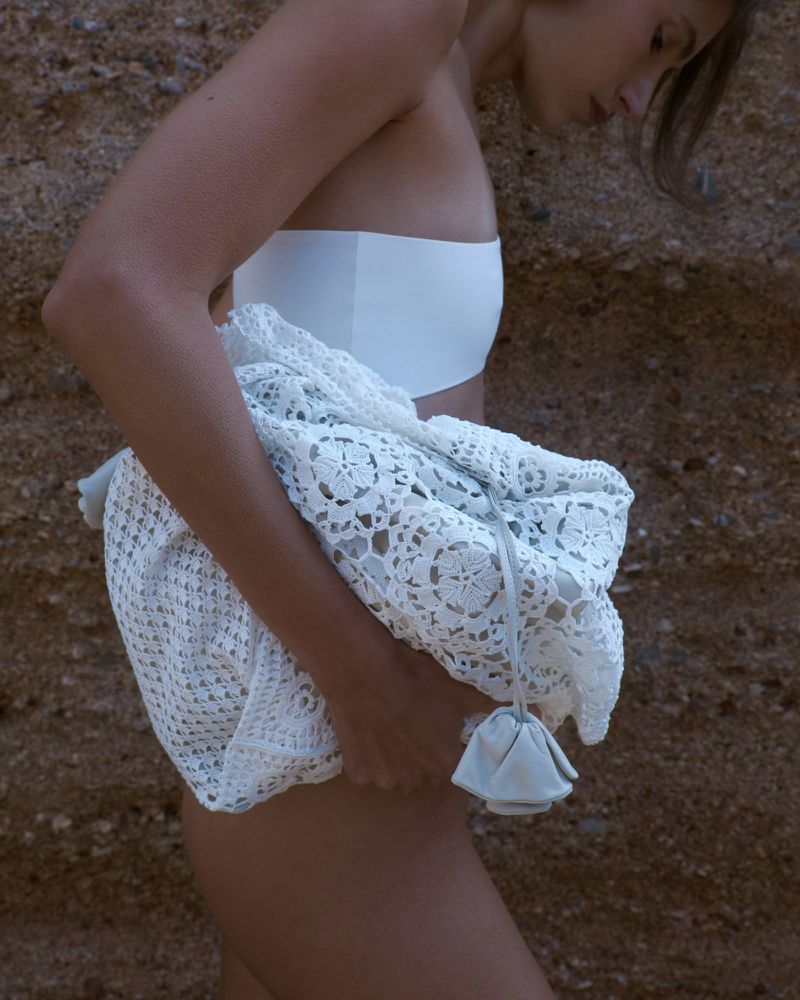 Woman in white swimwear holds a white lace bag near a sandy wall in an outdoor setting, emphasizing fashion and natural textures.