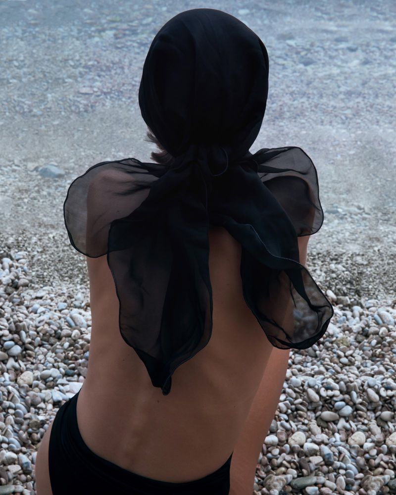 Back view of a person in a black headscarf and swimsuit sitting on a pebble beach with a view of a calm body of water.