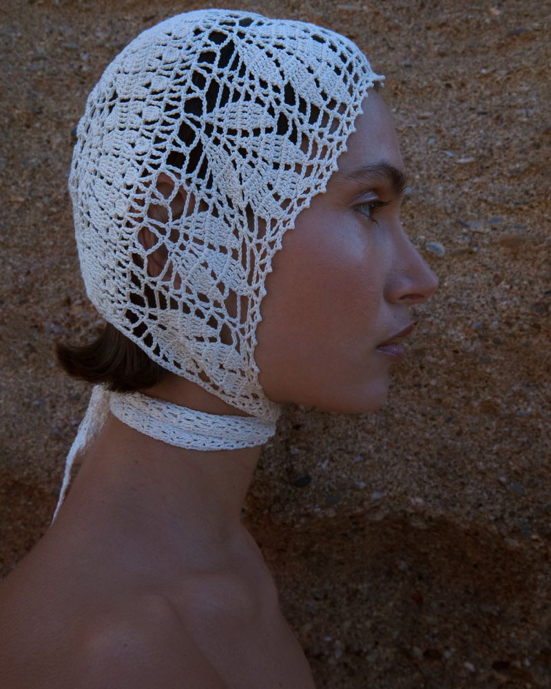 Profile shot of a woman wearing a white crochet bonnet with ties, set against a textured tan background, capturing a blend of fashion and craftsmanship.