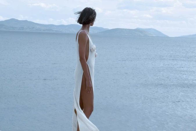 A woman in a white dress stands near the ocean with mountains in the background, looking out over the water on a windy day.