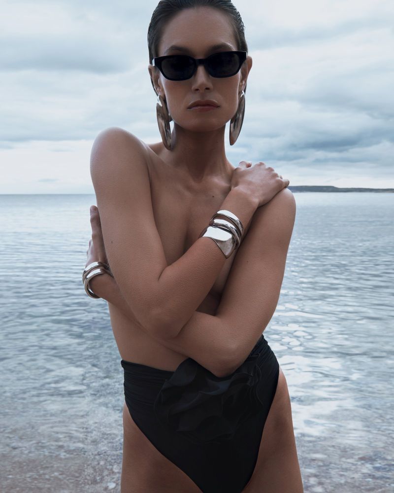 Adult woman in black sunglasses, wearing large earrings and silver cuffs, posing against a water and sky background with a serious expression.