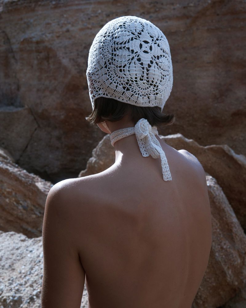 Back view of an adult female wearing a white crocheted head covering with tie strings against a tan rock background in a sunlit outdoor environment.
