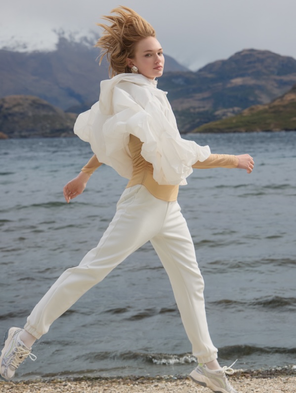 A young woman in white clothing jumps on a beach with water and mountains visible in the background on a cloudy day.