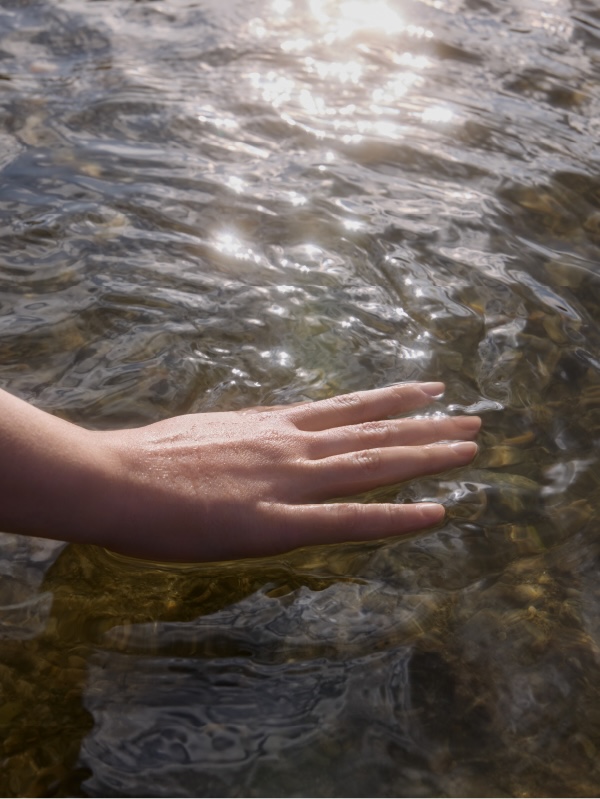 Close-up shot of an outstretched human hand gently touching the glistening surface of shallow water, sunlight creating sparkling reflections.