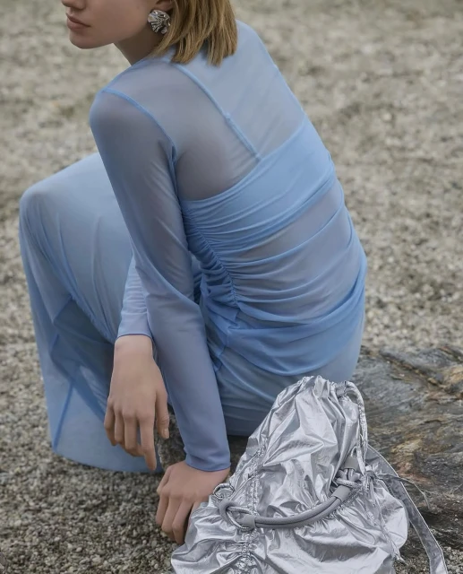 A fair-skinned young adult woman sits with a silver metallic bag on a rocky surface, wearing a light blue long-sleeve dress and earrings.