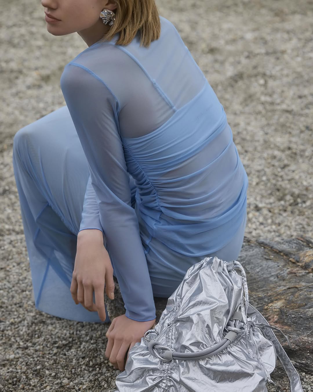 A young woman poses in a long blue dress next to a metallic silver bag on a gravelly surface, creating a minimalist fashion image.