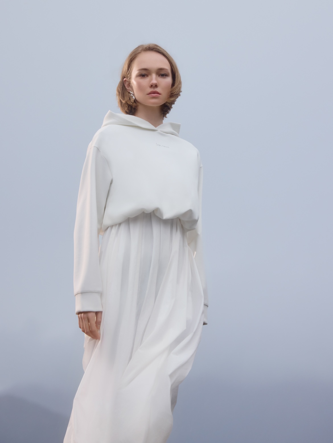 Blond woman in white hoodie and long skirt stands in a minimalist setting, looking at the camera with a neutral expression and soft lighting.