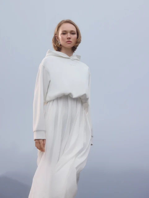 Blond woman in white hoodie and long skirt stands in a minimalist setting, looking at the camera with a neutral expression and soft lighting.