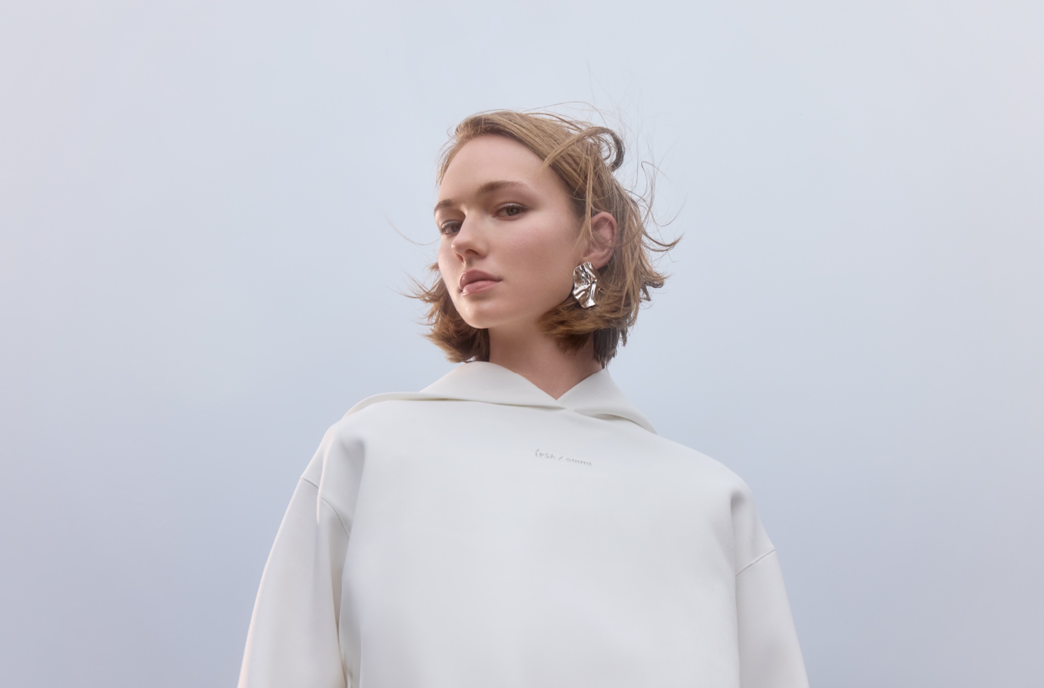Woman in white hoodie and statement earrings poses against a pale background in a fashion shot; casual yet stylish.
