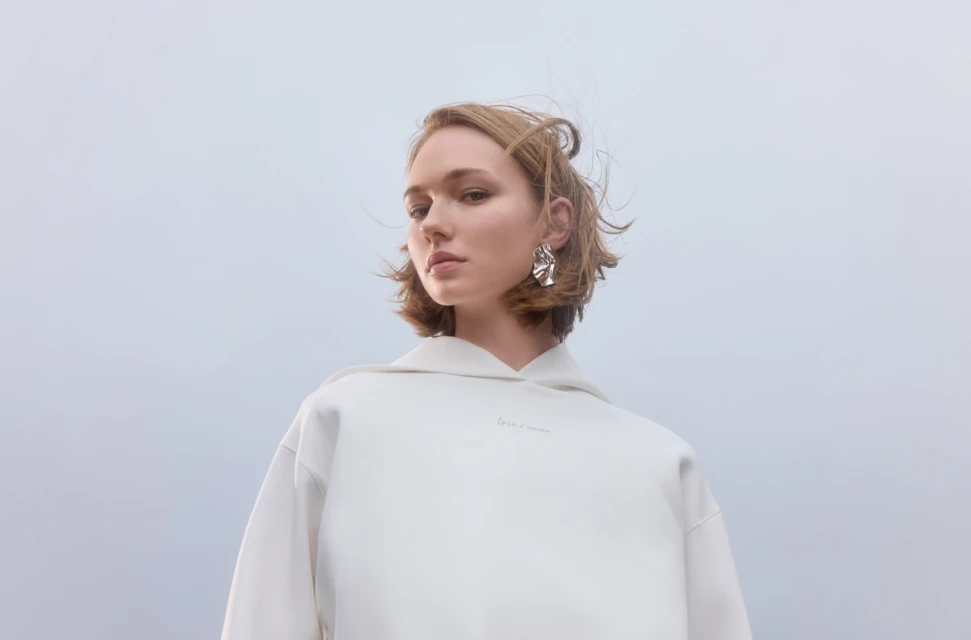 Woman in white hoodie and statement earrings poses against a pale background in a fashion shot; casual yet stylish.