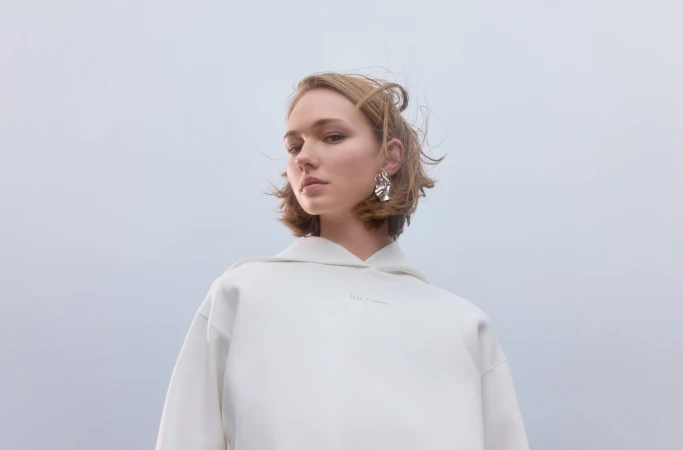 Woman in white hoodie and statement earrings poses against a pale background in a fashion shot; casual yet stylish.
