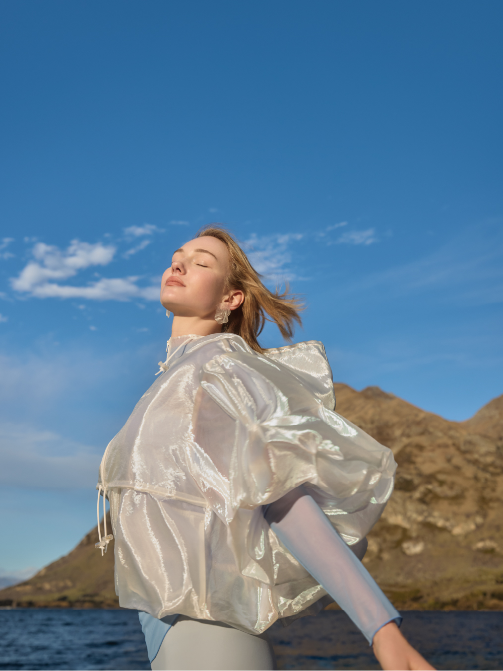 Adult woman with closed eyes in a puffy translucent jacket and blue sleeves poses in front of a mountain and ocean backdrop on a sunny day.