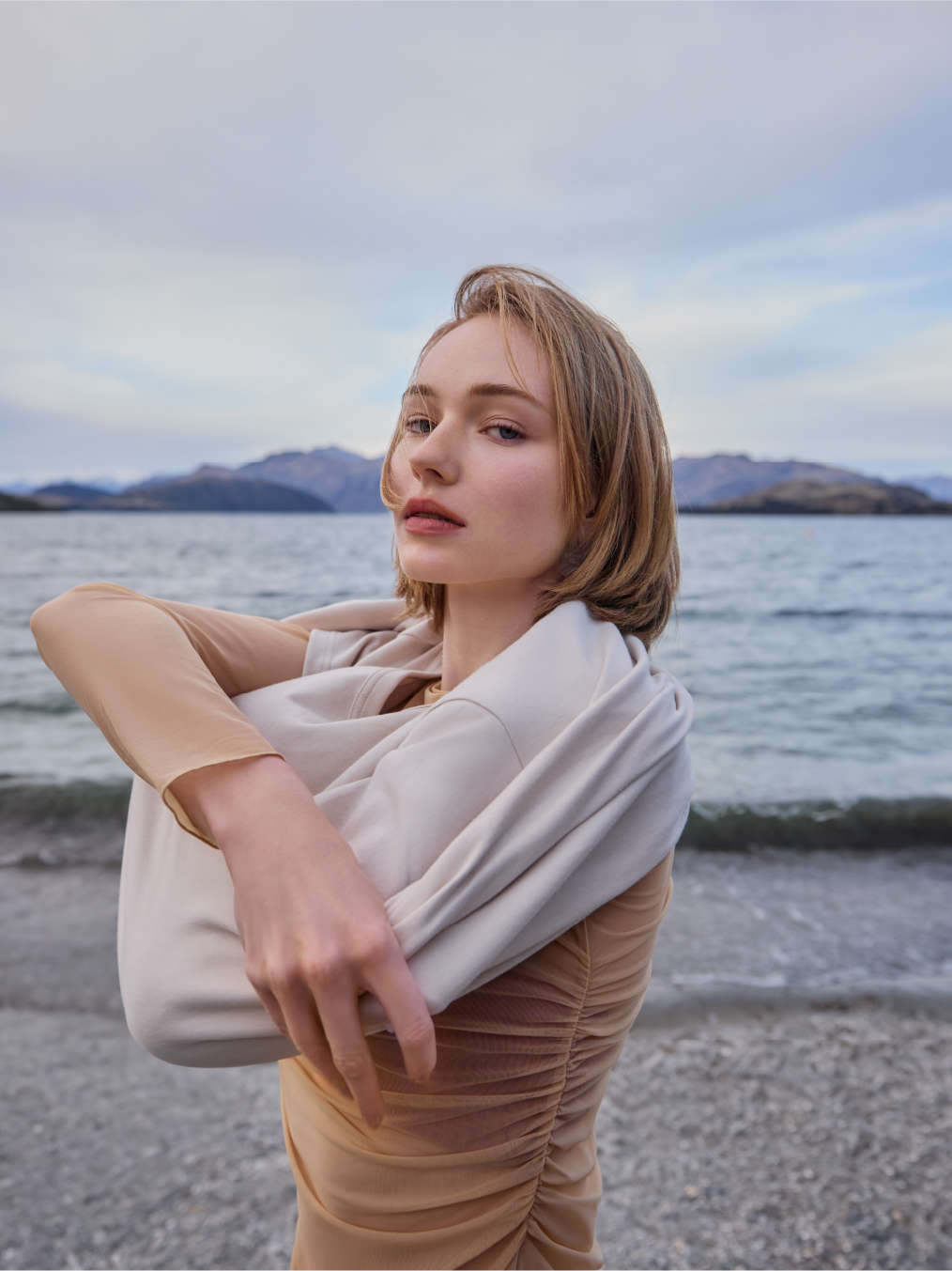 Young woman in dress poses on pebble beach with mountains and cloudy sky behind her in medium shot. Neutral tones, serene mood.
