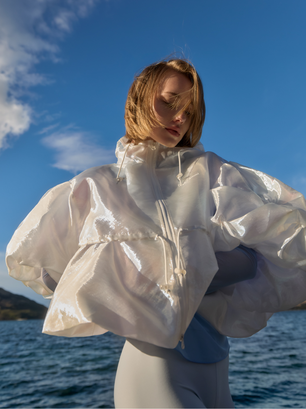 Woman with windswept hair poses in a glossy white translucent jacket with puffy sleeves over a bodysuit against a blue sky and sea backdrop.