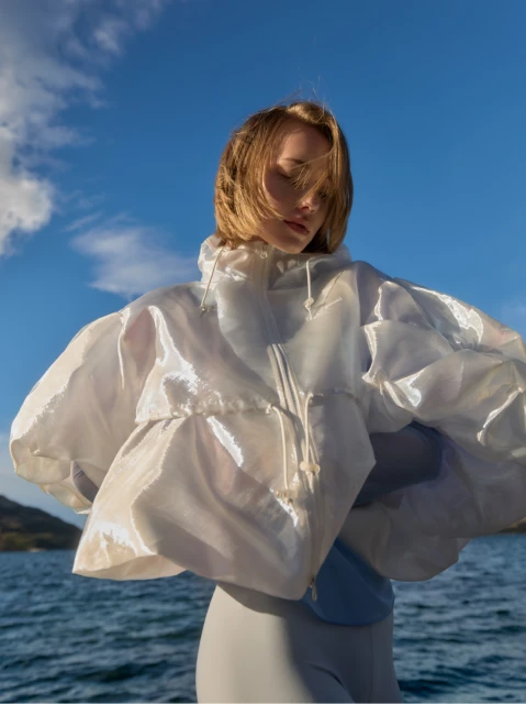 Woman with windswept hair poses in a glossy white translucent jacket with puffy sleeves over a bodysuit against a blue sky and sea backdrop.