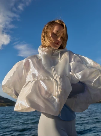 Woman with windswept hair poses in a glossy white translucent jacket with puffy sleeves over a bodysuit against a blue sky and sea backdrop.